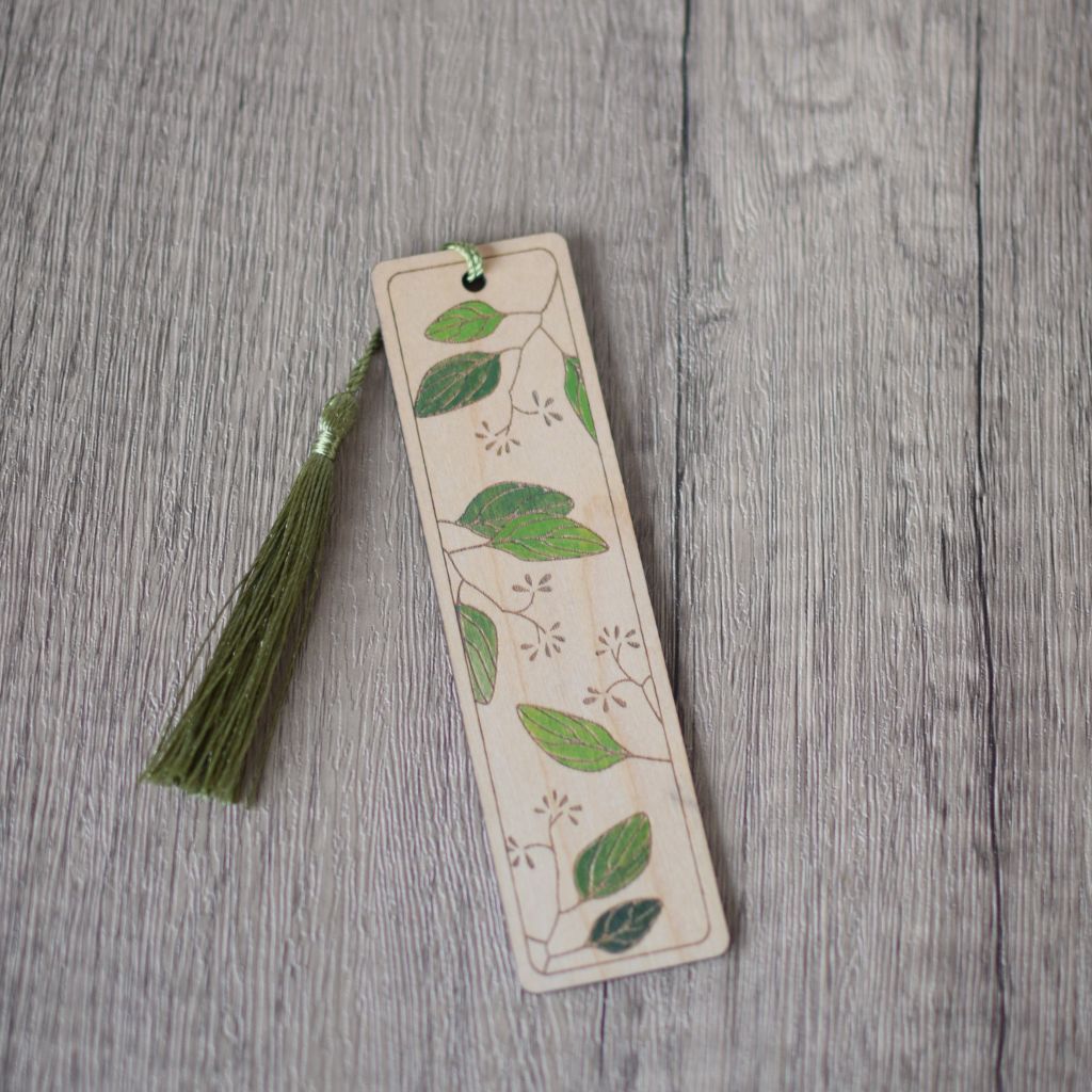 A wooden laser engraved bookmark with a leaves design. The leaves are then hand painted with shades of green. The bookmark is fitted moss green silky tassel