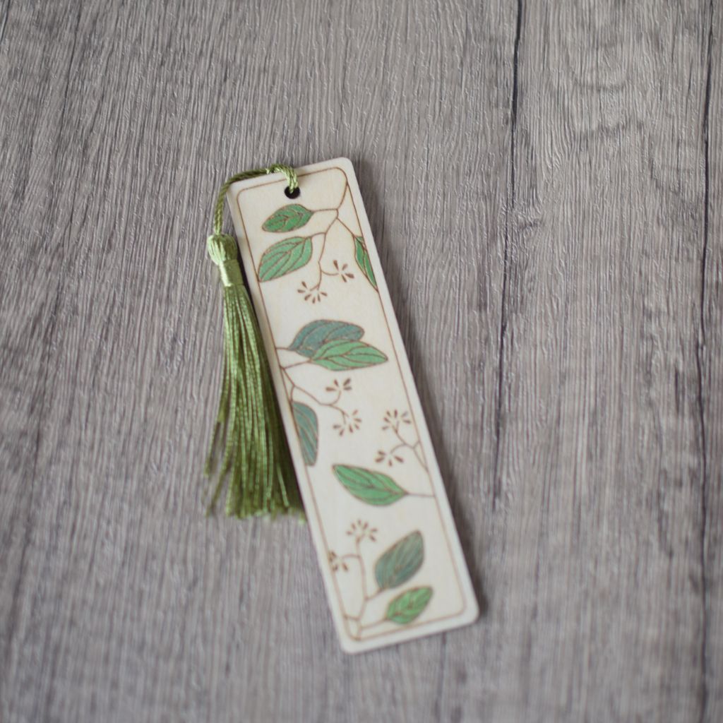 A single wooden laser engraved bookmark with a leaves design. The leaves are hand painted in shades of metallic green. The bookmark is fitted with a moss green silky tassel